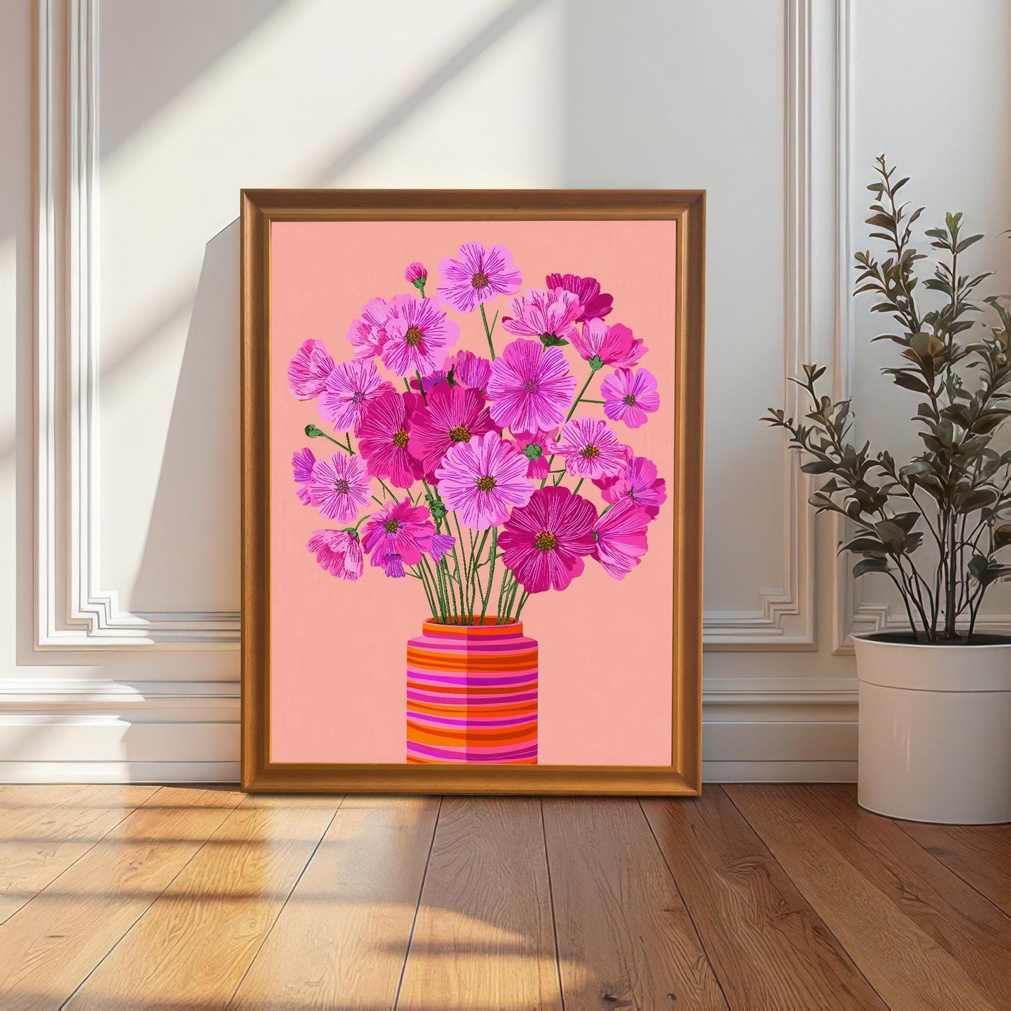 The pink Cosmos flower print in an orange/gold frame, leaning on the floor against a white paneled wall next to a tall potted plant.