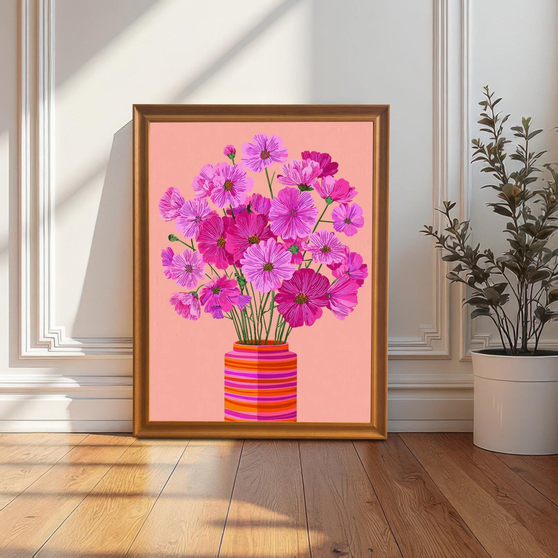 The pink Cosmos flower print in an orange/gold frame, leaning on the floor against a white paneled wall next to a tall potted plant.