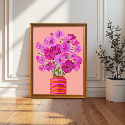 The pink Cosmos flower print in an orange/gold frame, leaning on the floor against a white paneled wall next to a tall potted plant.