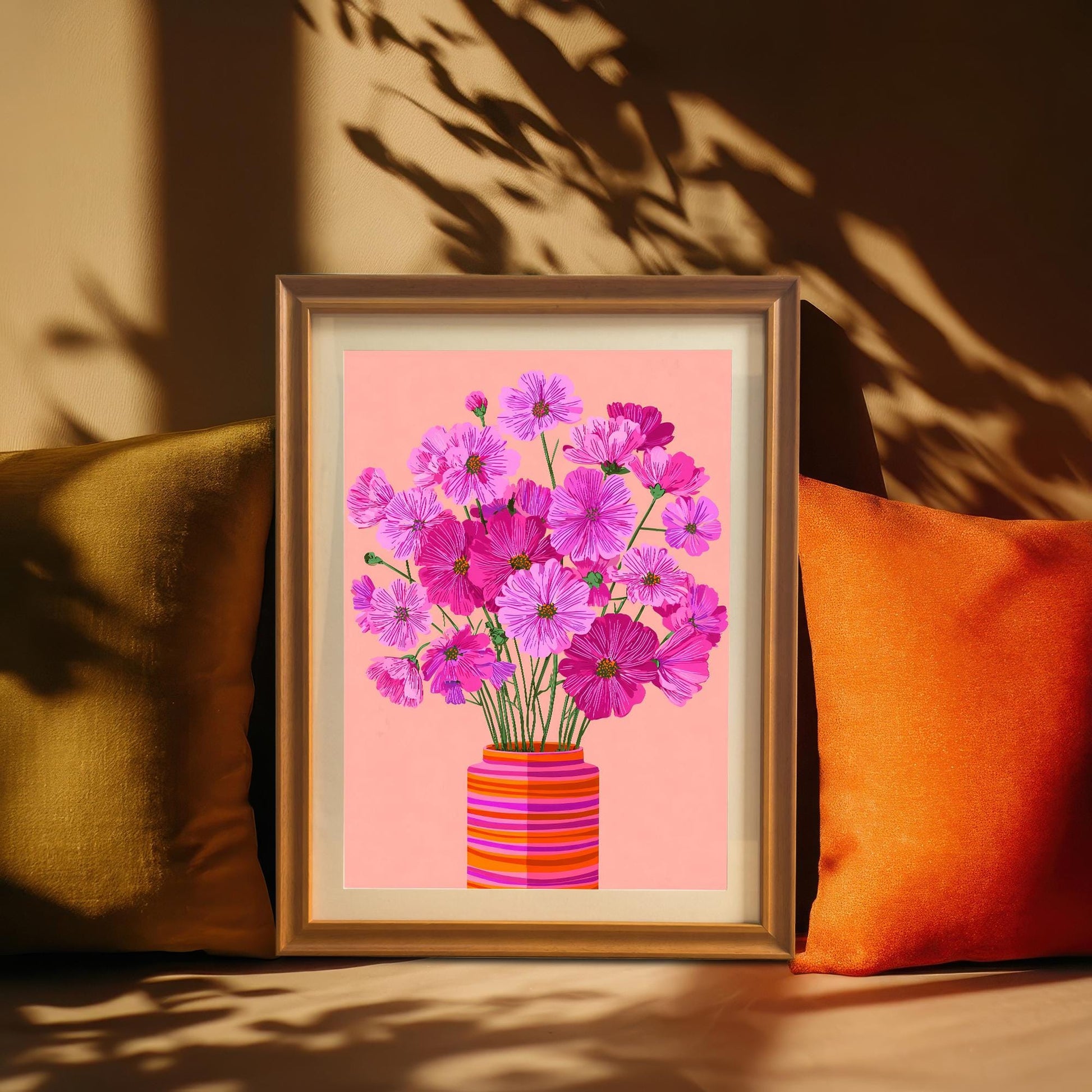 The framed print of pink Cosmos flowers in a gold frame, leaning between an olive green and an orange pillow, with dramatic plant shadows cast on the beige wall.