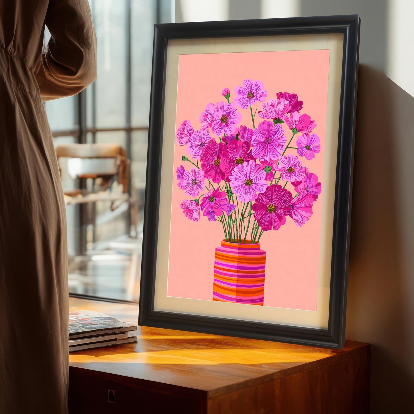 Vertical image of the Cosmos flower print in a black frame with a mount (matting), leaning on a wooden cabinet near a window. A person in a long beige dress stands to the left.