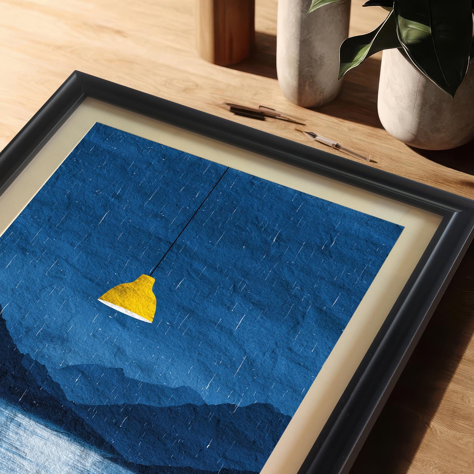 A close-up, high-angle view of the dark blue rainy landscape art print in a black frame with a light mat/mount, resting on a wooden desk or table. The bright yellow lamp contrasts strongly with the dark blue mountains and water.