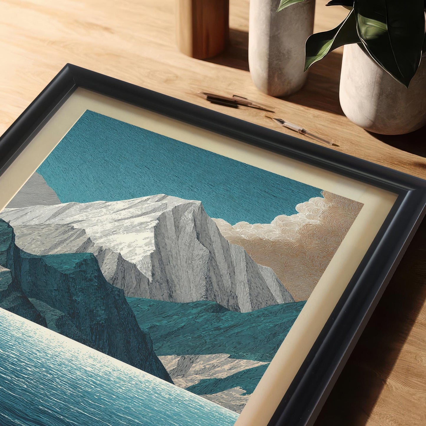 A close-up, high-angle view of the mountain and lake art print in a black frame with a light mat/mount, resting on a wooden desk or table. Art supplies (pencils) and potted plants are visible in the blurred background.