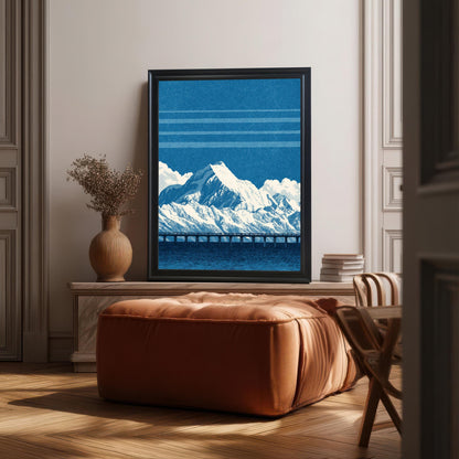 The blue mountain and bridge art print in a black frame, resting on a small, light-colored console table or mantelpiece, positioned above a large, cushioned, rust-colored ottoman/pouf. A decorative vase and stack of books are also visible, with sunlight creating deep shadows on the herringbone floor.