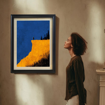 A woman in a striped shirt looks up at the abstract yellow cliff print in a Black frame with a mount/mat hanging on a textured tan wall. The print features a figure on a cliff edge against a deep blue sky.