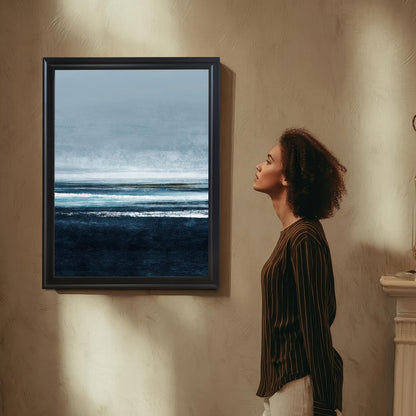 Abstract seascape print with dark navy blue waves/water at the bottom, layered white and light blue lines representing the horizon and sky, in a Black frame hanging on a textured tan wall. A woman in a striped shirt looks up at the artwork.