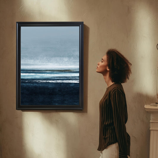 Abstract seascape print with dark navy blue waves/water at the bottom, layered white and light blue lines representing the horizon and sky, in a Black frame hanging on a textured tan wall. A woman in a striped shirt looks up at the artwork.