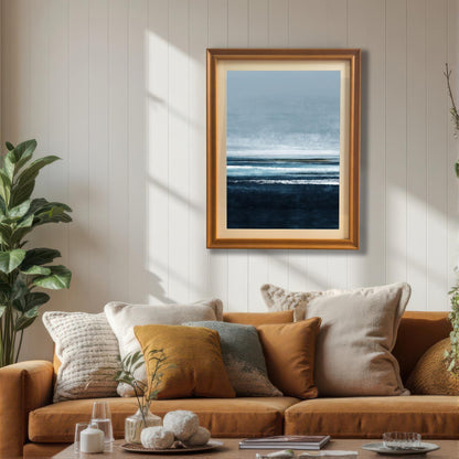 Framed abstract seascape print in an Orange/light brown wood frame hanging above a tan sofa with pillows, in a sunlit living room with a houseplant. The print features deep navy, light blue, and white brushstrokes.
