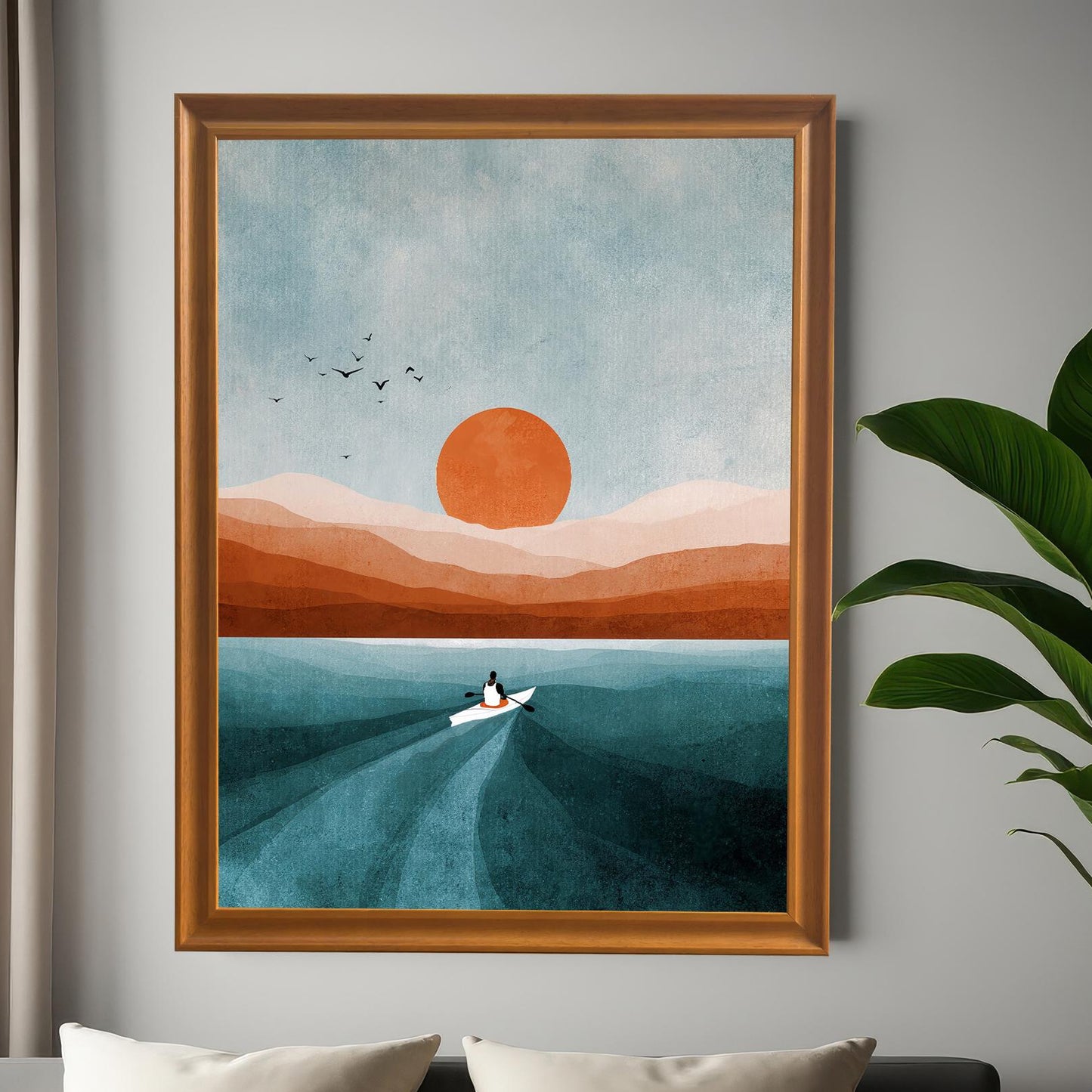 Large, framed wall art of a boat on water at sunset/sunrise, mounted on a gray wall above a modern bed with white pillows. The frame is brown wood. A large green potted plant is on the right. Shows the print in a bedroom setting.