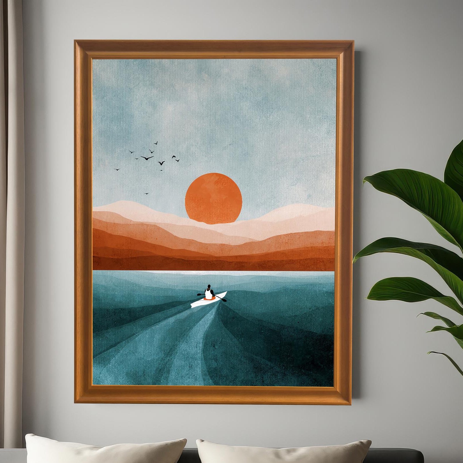 Large, framed wall art of a boat on water at sunset/sunrise, mounted on a gray wall above a modern bed with white pillows. The frame is brown wood. A large green potted plant is on the right. Shows the print in a bedroom setting.