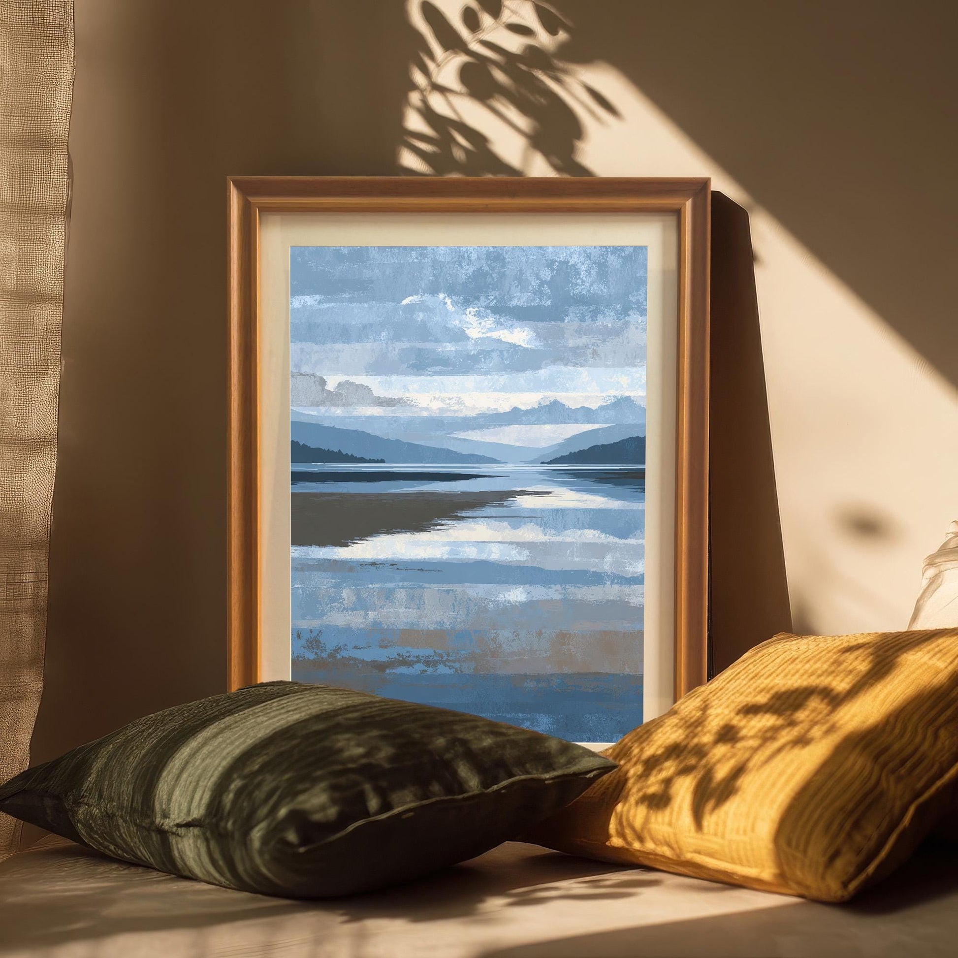 Abstract landscape art print framed in Orange wood with a light-colored mount, propped up between dark green and yellow throw pillows on a couch, with sunlight and shadows cast above.