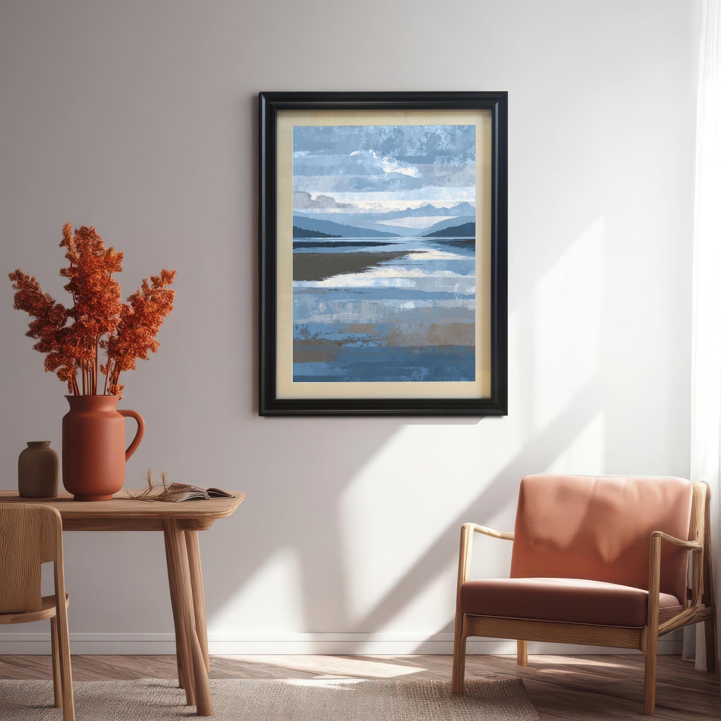 Abstract blue and brown landscape art print framed in black wood with a light mount, hanging on a white wall in a sunlit dining area with a wooden table, terracotta vase, and an upholstered wooden armchair.