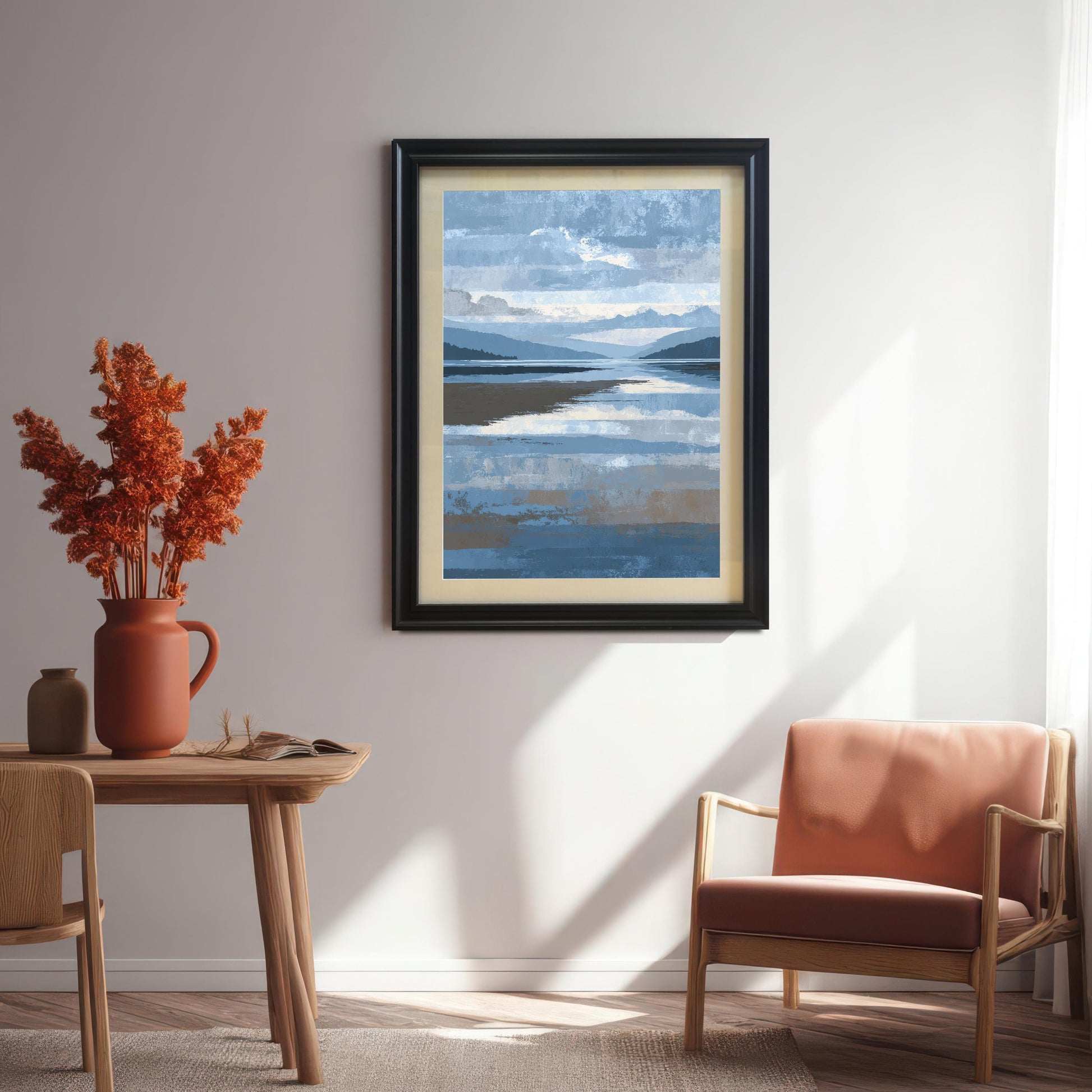 Abstract blue and brown landscape art print framed in black wood with a light mount, hanging on a white wall in a sunlit dining area with a wooden table, terracotta vase, and an upholstered wooden armchair.