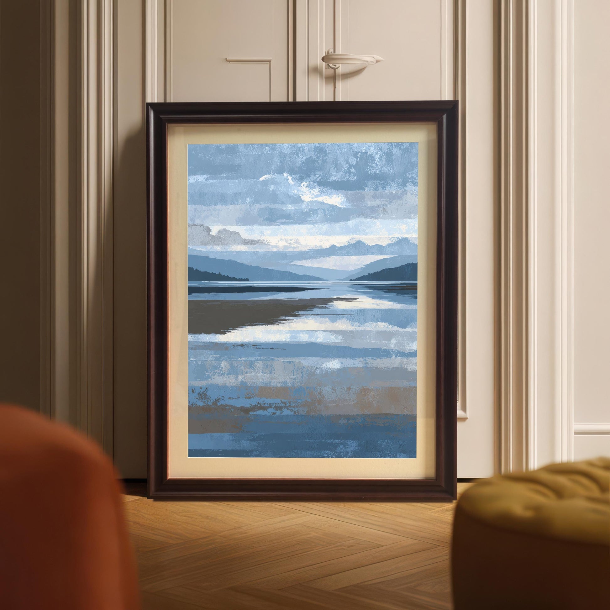 Abstract blue and brown landscape art print framed in dark Coffee wood with a light mount, leaning against a white panel door on a wooden floor, partially visible behind an orange chair and yellow ottoman.