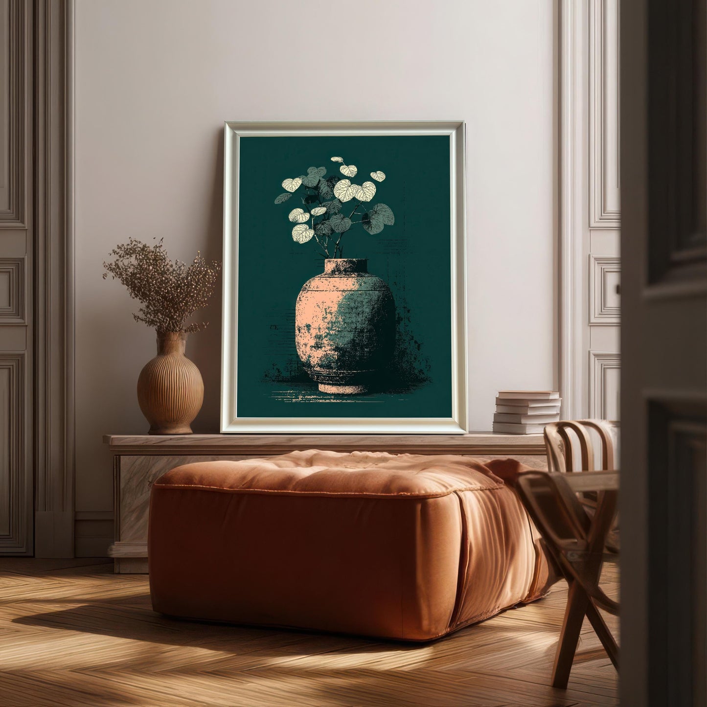 A framed art print of a distressed vase with pale leaves on a dark teal background, leaning against a pale, paneled wall above a square, burnt-orange leather ottoman. The frame is White (or light silver).