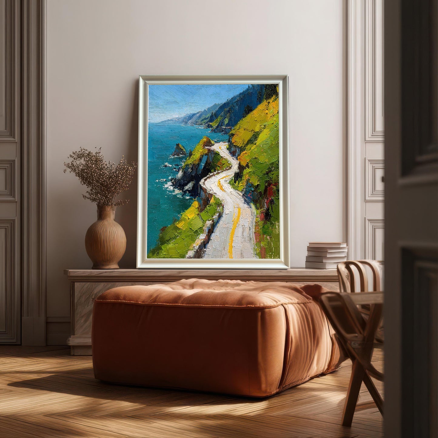 A framed print of a vibrant oil painting of a winding coastal road, leaning against a pale, paneled wall above a square, burnt-orange leather ottoman. The frame is White (or light silver).
