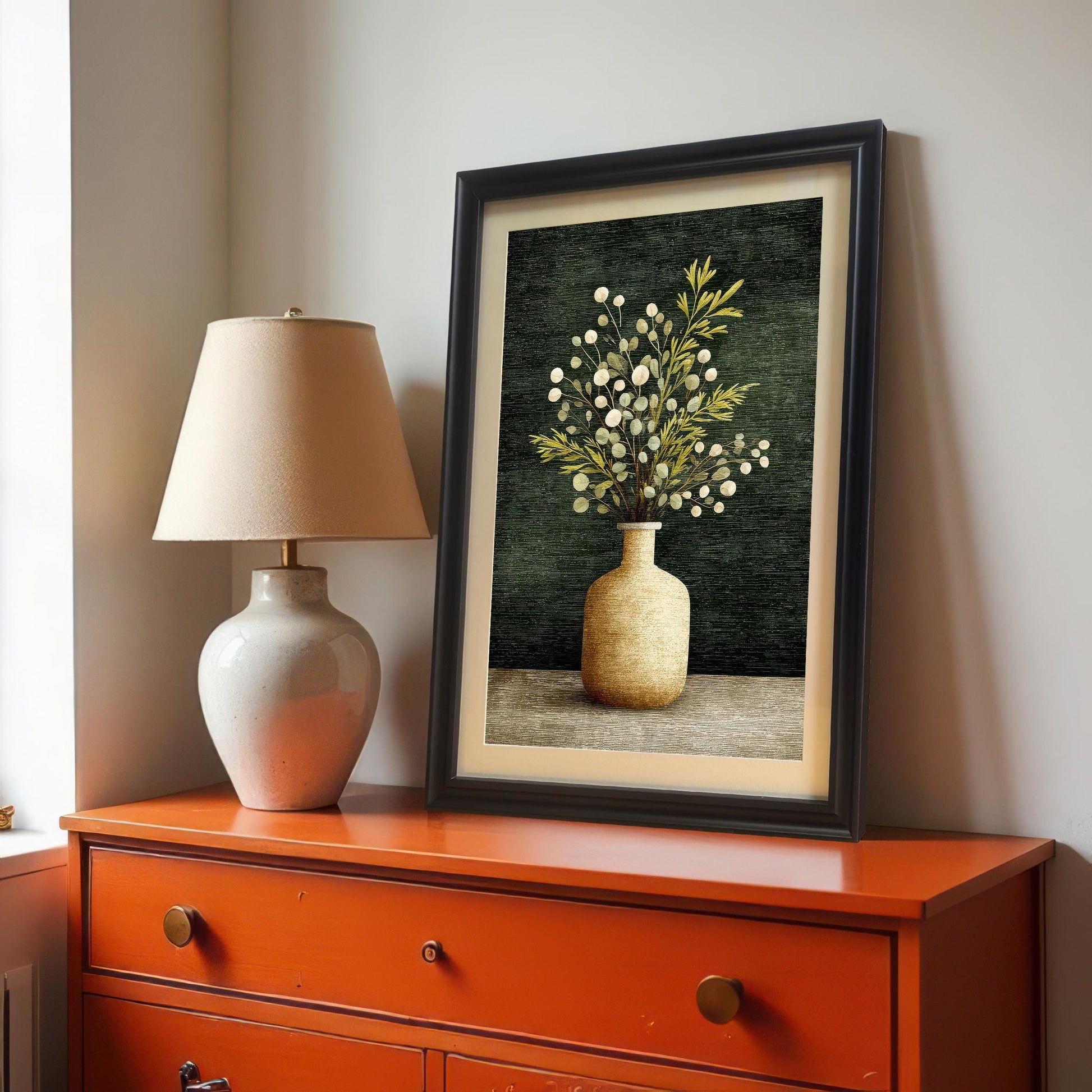 A framed art print of a vase with foliage, leaning on an Orange/Red dresser. The frame is a Black wood with a light border/mount. A cream-colored table lamp is next to the print.