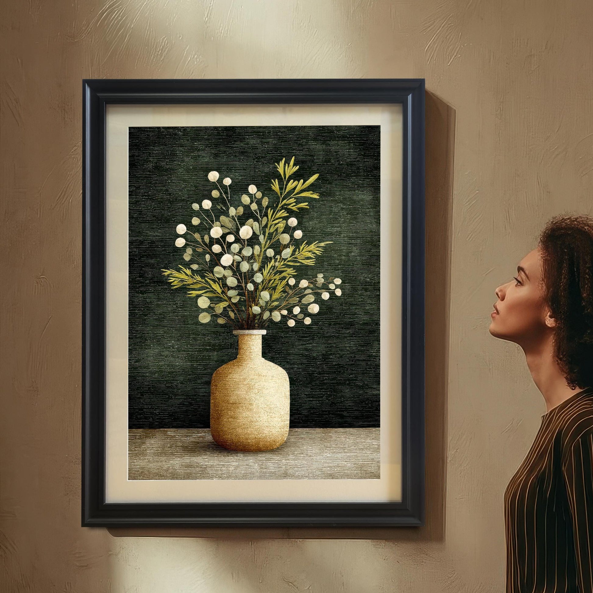 A woman with curly hair is standing and looking up at a framed art print of a vase with foliage, which is hanging on a textured beige wall. The frame is a Black wood with a light border/mount.