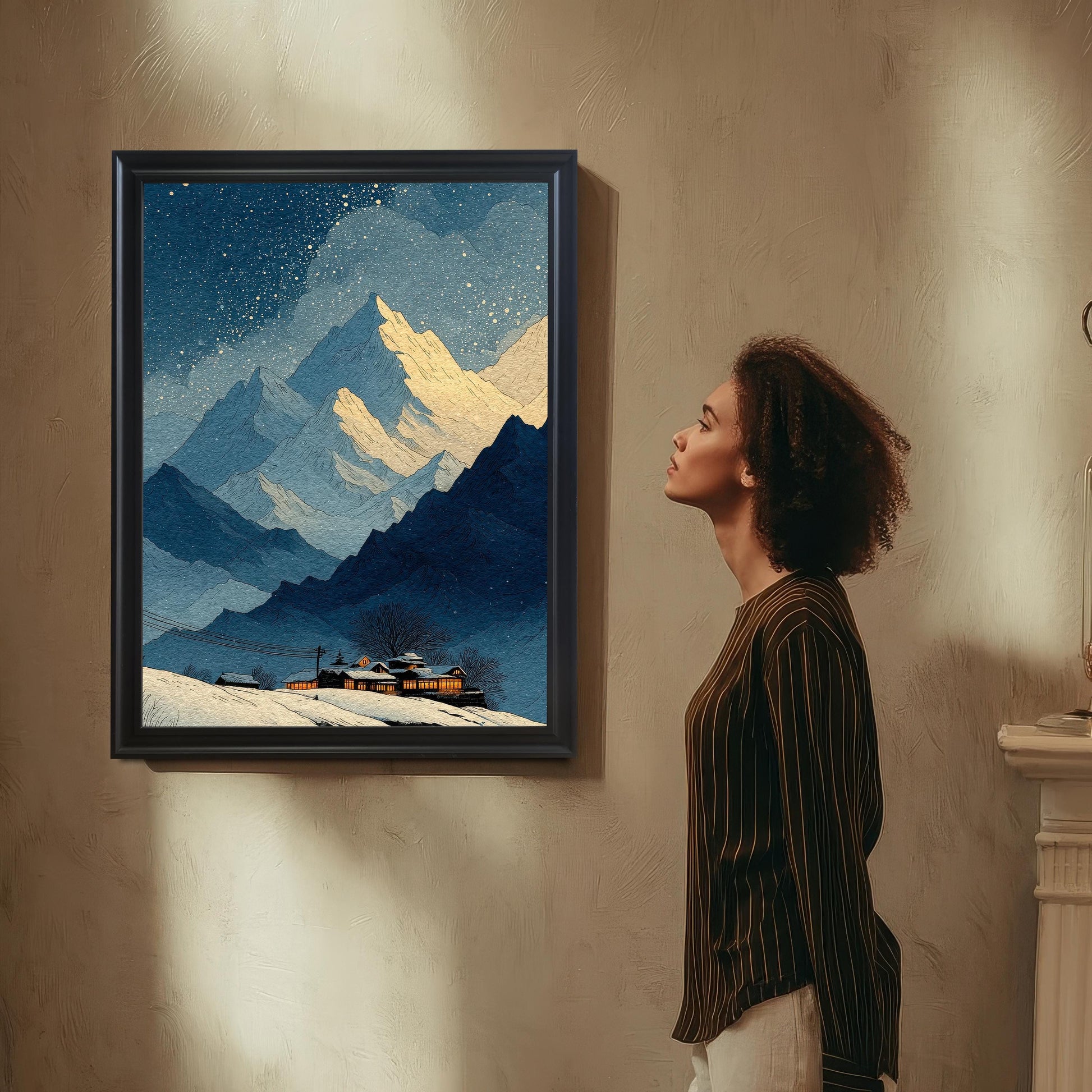 A woman with curly hair is standing and looking up at a framed print of a snowy mountain scene. The frame is Black with a light border/mount. The wall is textured beige.
