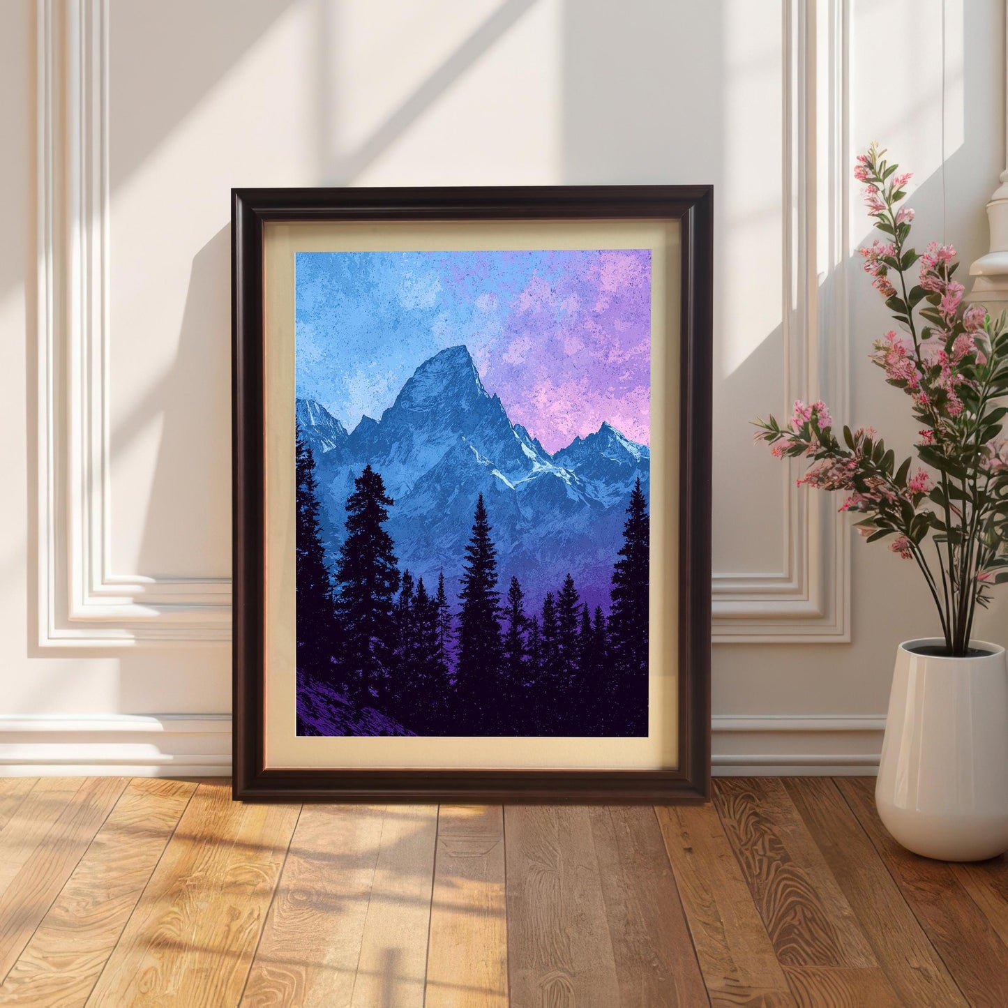 A framed mountain art print with a Coffee Frame & Mount (dark brown frame with light matting) is propped on a hardwood floor against a white paneled wall. Sunlight casts strong shadows, and a potted pink flowering plant is visible on the right.