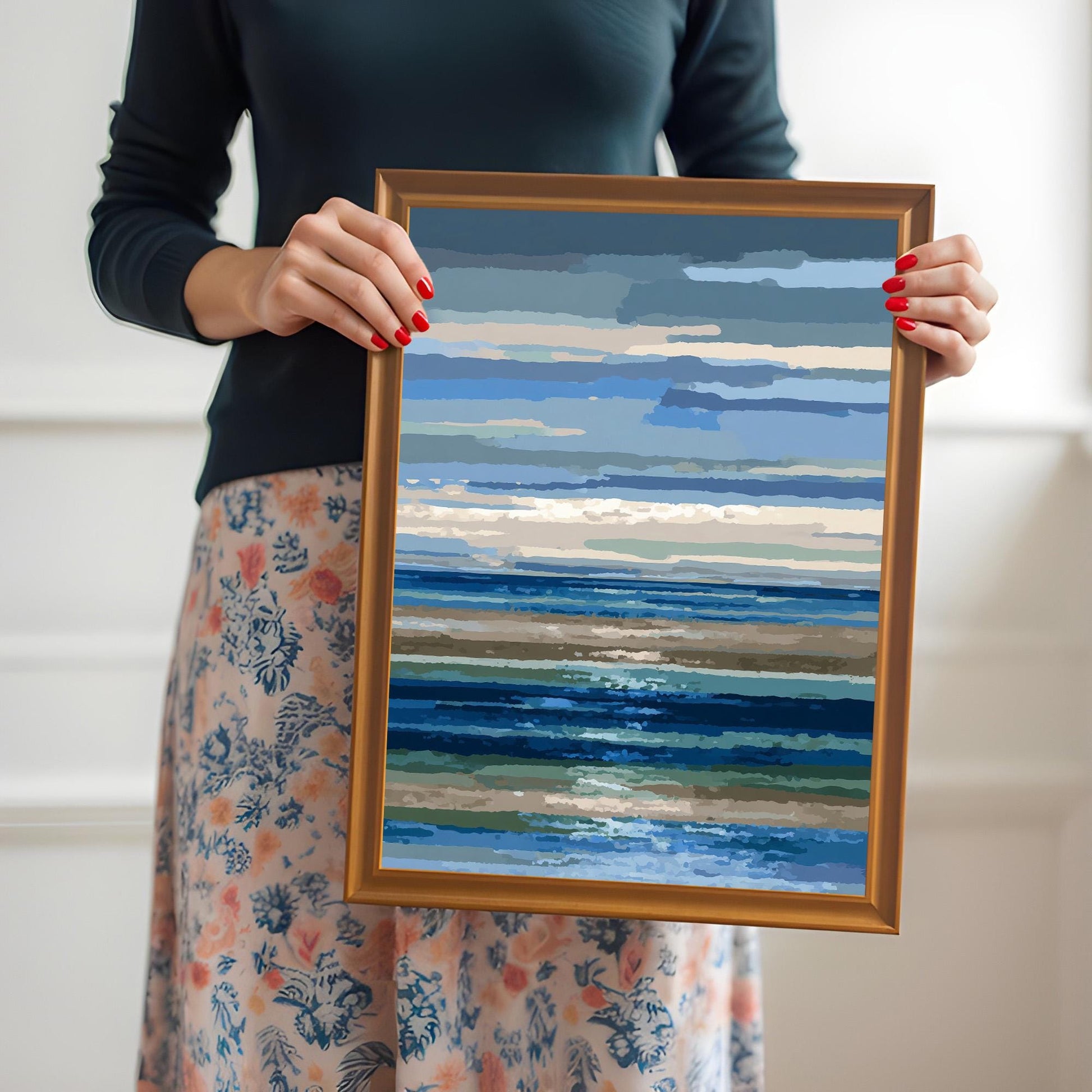 A woman in a dark shirt and floral skirt holding the abstract blue and brown striped seascape print in a gold/orange wood frame.