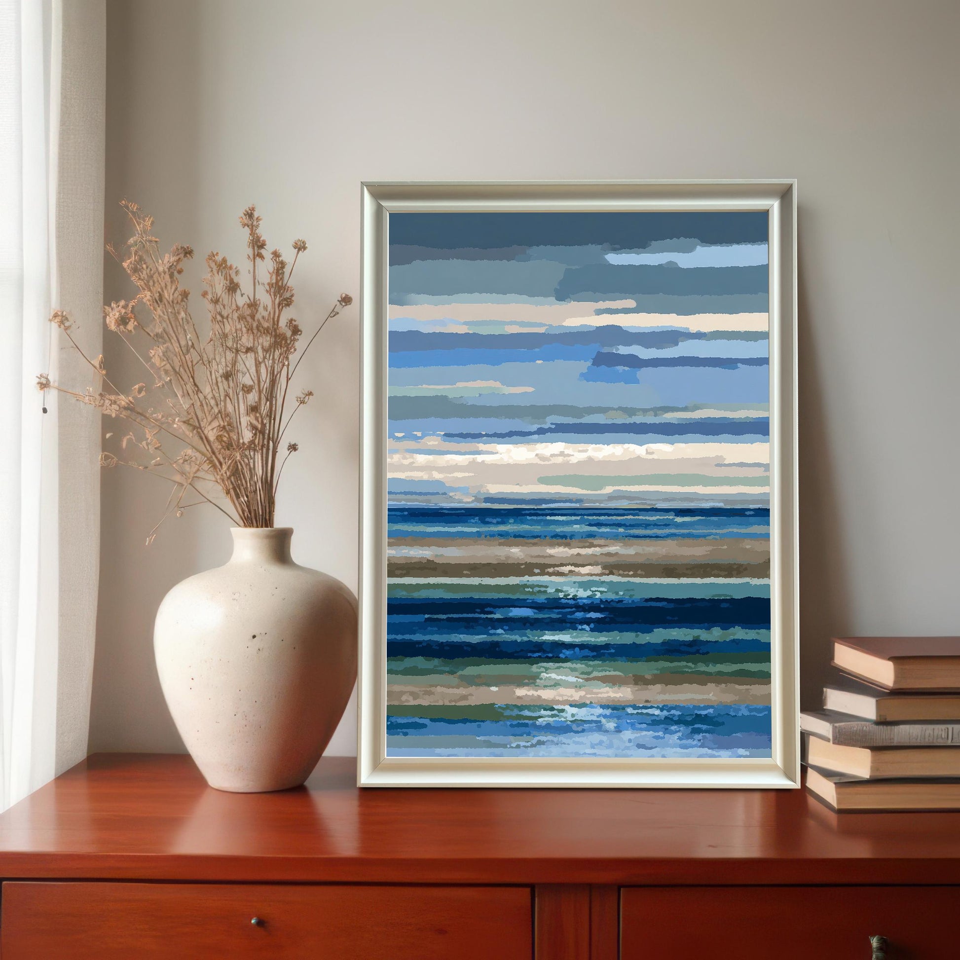 Abstract blue and brown striped seascape art print in a white frame, displayed on a wooden dresser next to a ceramic vase with dried flowers and a stack of books.