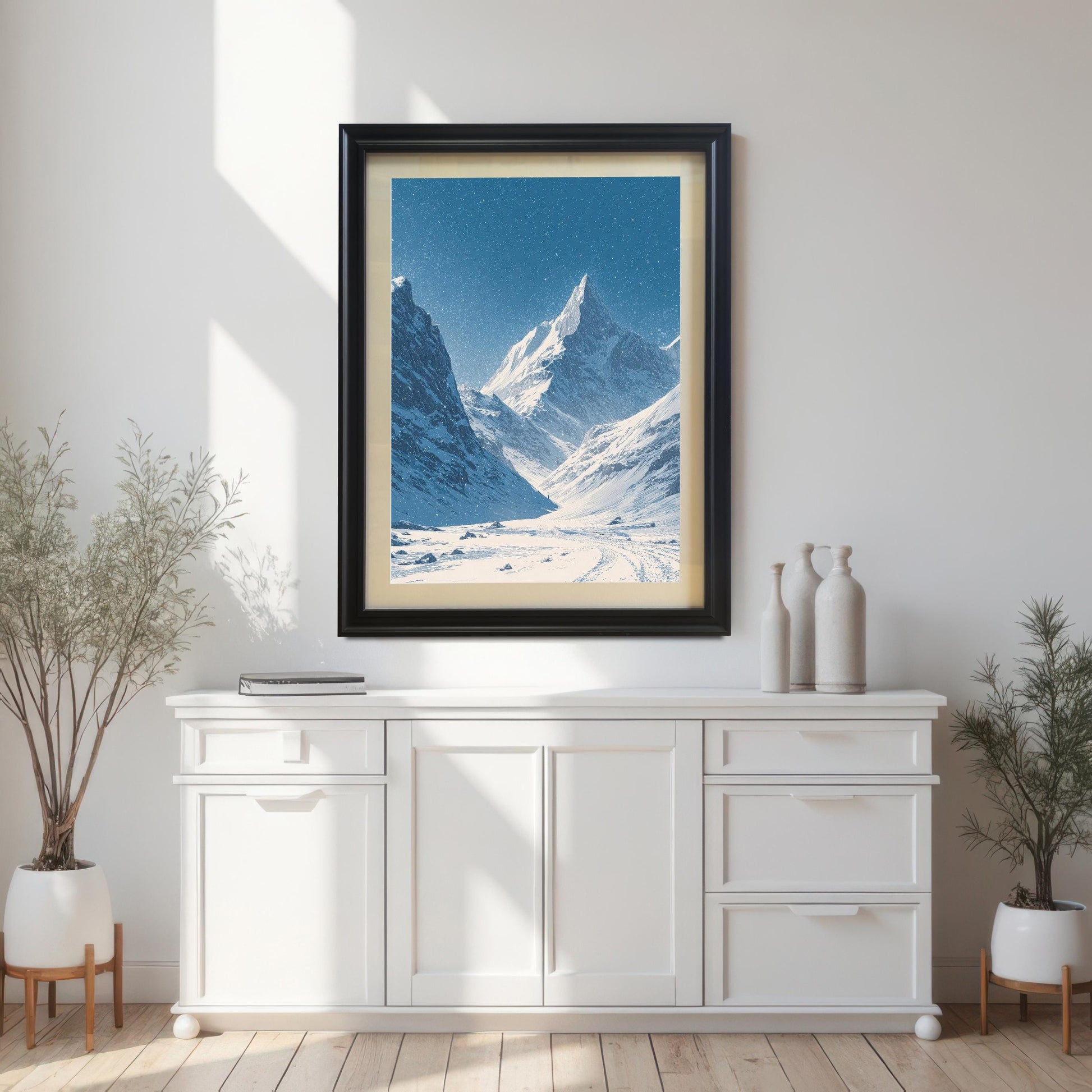 Snowy mountain valley print in a black wood frame with a light mount, hanging above a white console table in a bright room with potted plants.