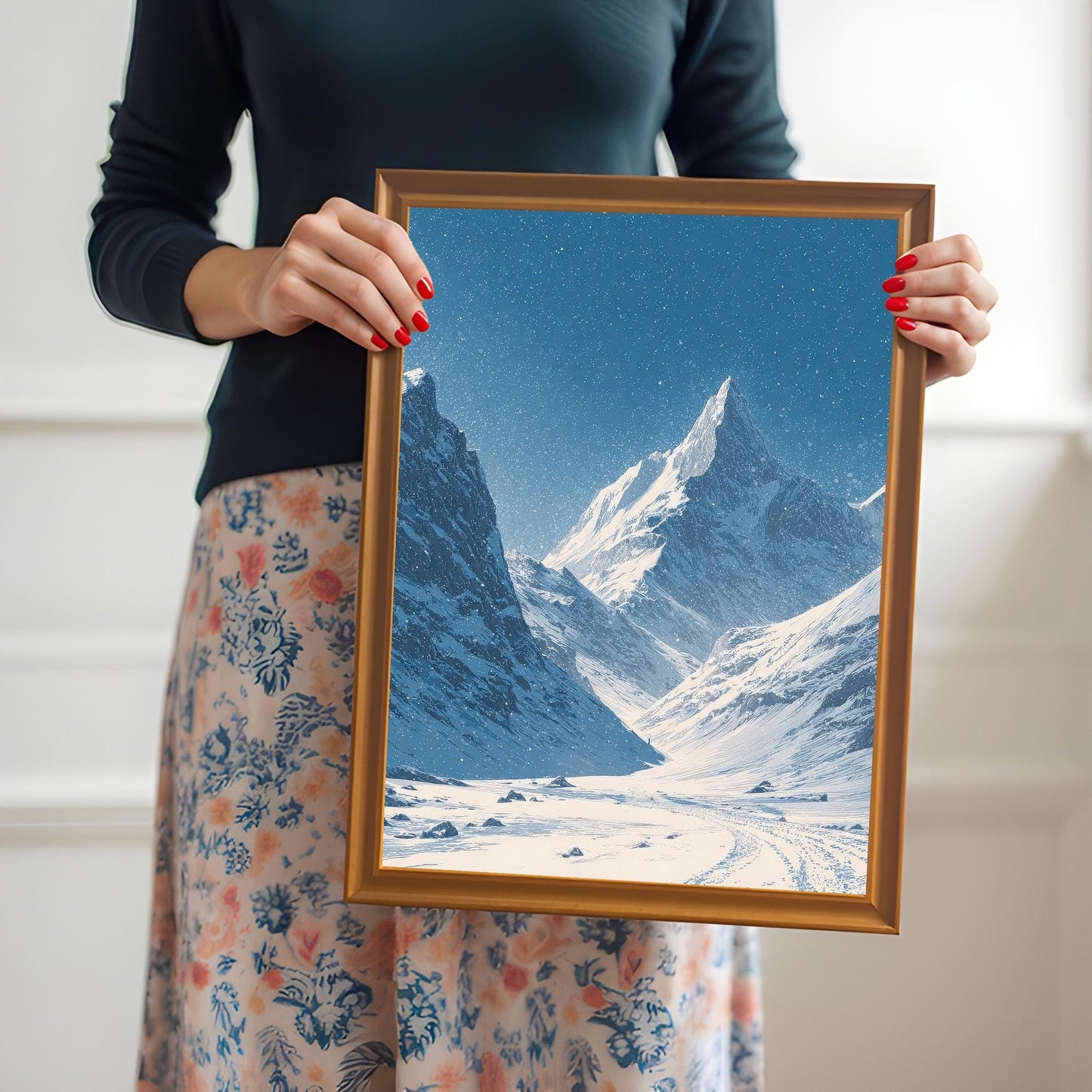 A woman in a dark shirt and floral skirt holding the snowy mountain valley print in a gold/orange wood frame.