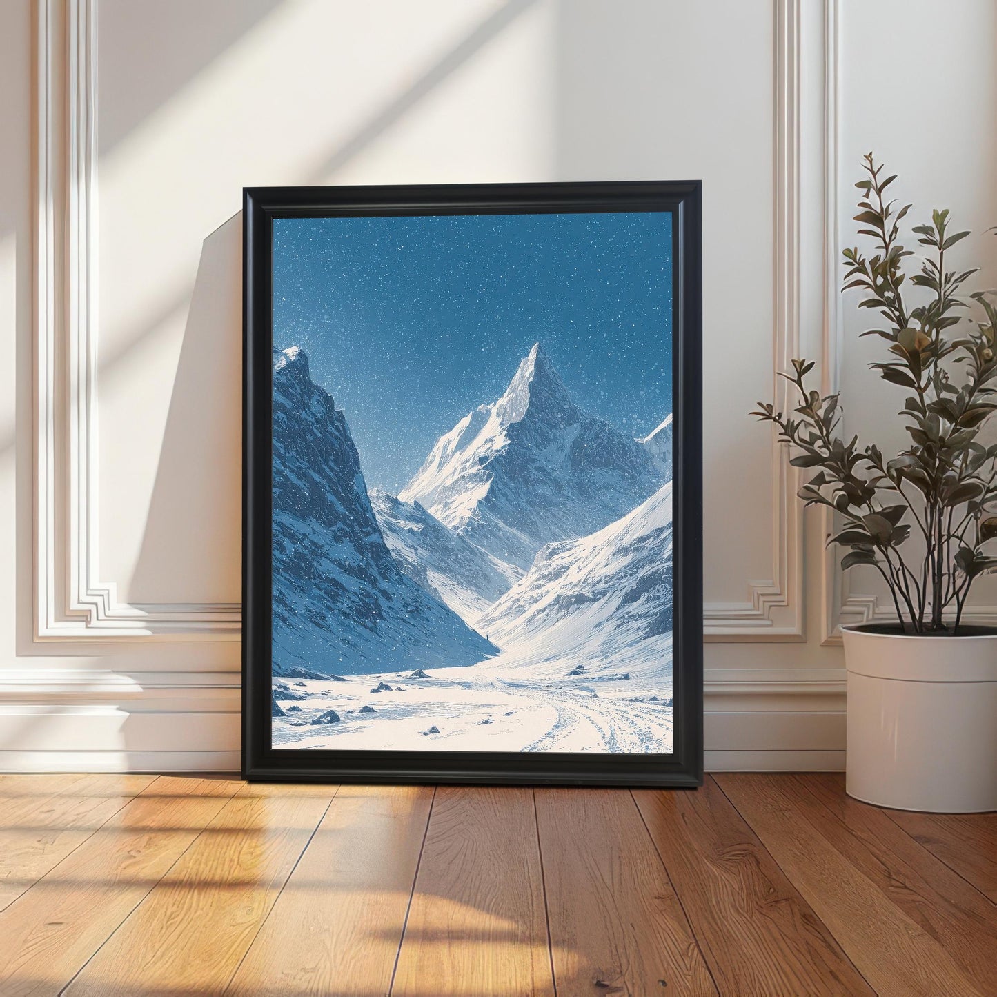 Snowy mountain valley print in a black frame, leaning on a wooden floor against a white paneled wall, with long shadows from sunlight and a potted plant nearby.