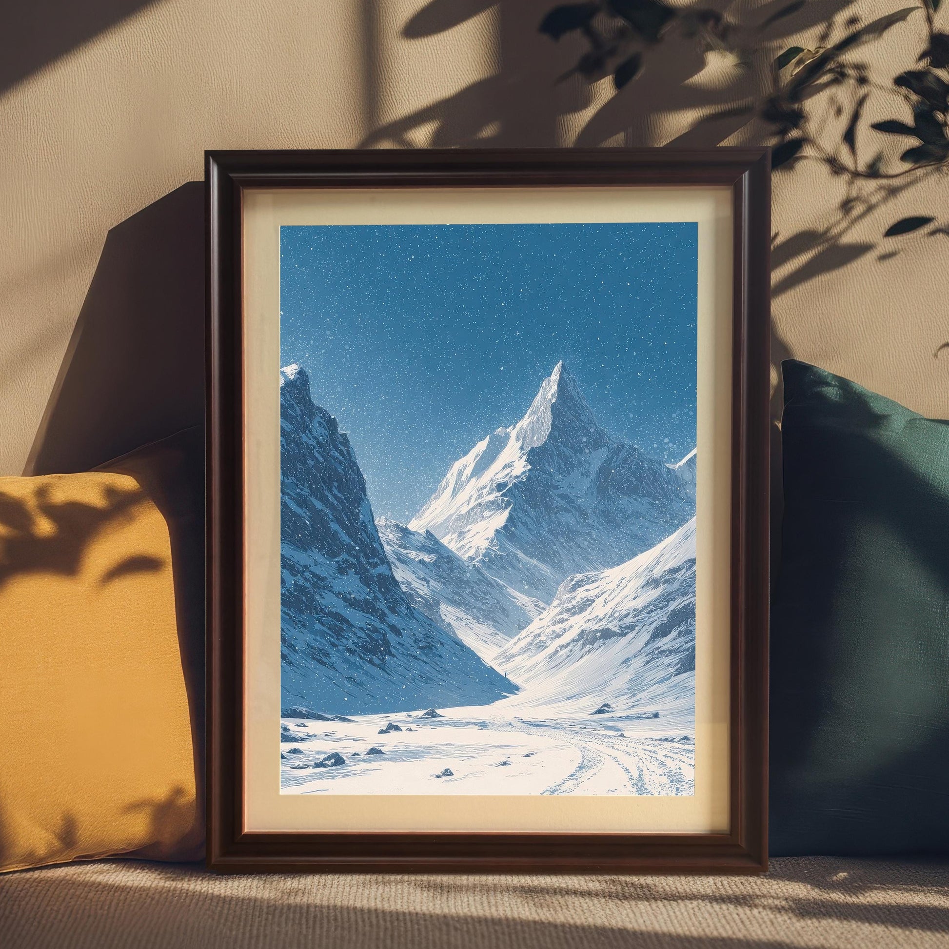 Snowy mountain valley print in a dark coffee frame with a mount, resting on a couch between yellow and teal pillows, with shadows of leaves cast on the wall.