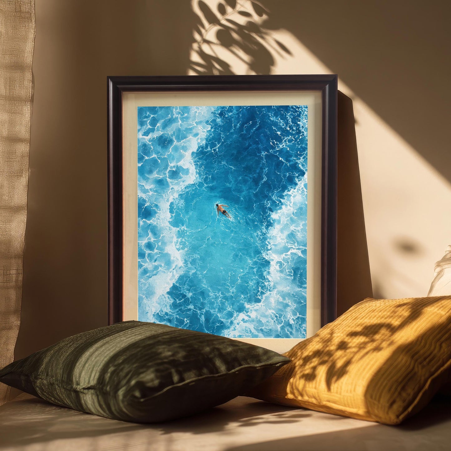 The framed ocean art print in a Coffee/dark brown frame with a mat (mount), leaning against a wall and surrounded by dark green and yellow cushions. Plant shadows are cast across the art and cushions.