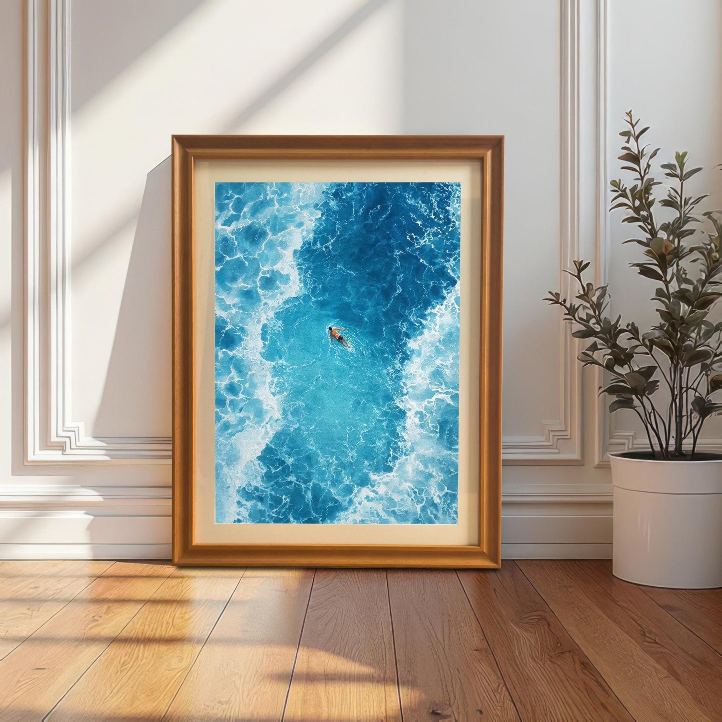 The framed ocean art print in an Orange/light brown frame with a mat (mount), leaning against a white wainscoted wall next to a large potted plant. Sunlight and shadows fall on the wall and wooden floor.