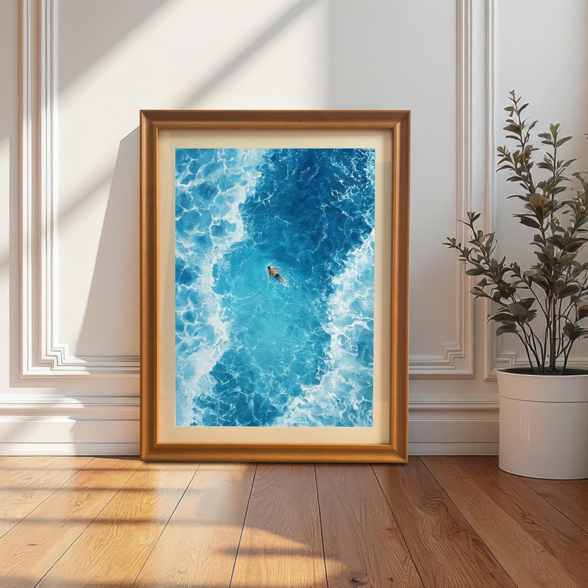 The framed ocean art print in an Orange/light brown frame with a mat (mount), leaning against a white wainscoted wall next to a large potted plant. Sunlight and shadows fall on the wall and wooden floor.