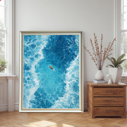 A large framed ocean art print in a White/silver frame with no mat, leaning on a wooden floor between two sunlit windows and a wooden chest of drawers with decorative vases and plants.