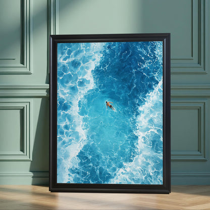 The framed ocean art print in a Black frame with no mat, leaning on a wooden floor against a wainscoted wall painted in a dark sage green color. Sunlight and plant shadows fall across the art and wall.