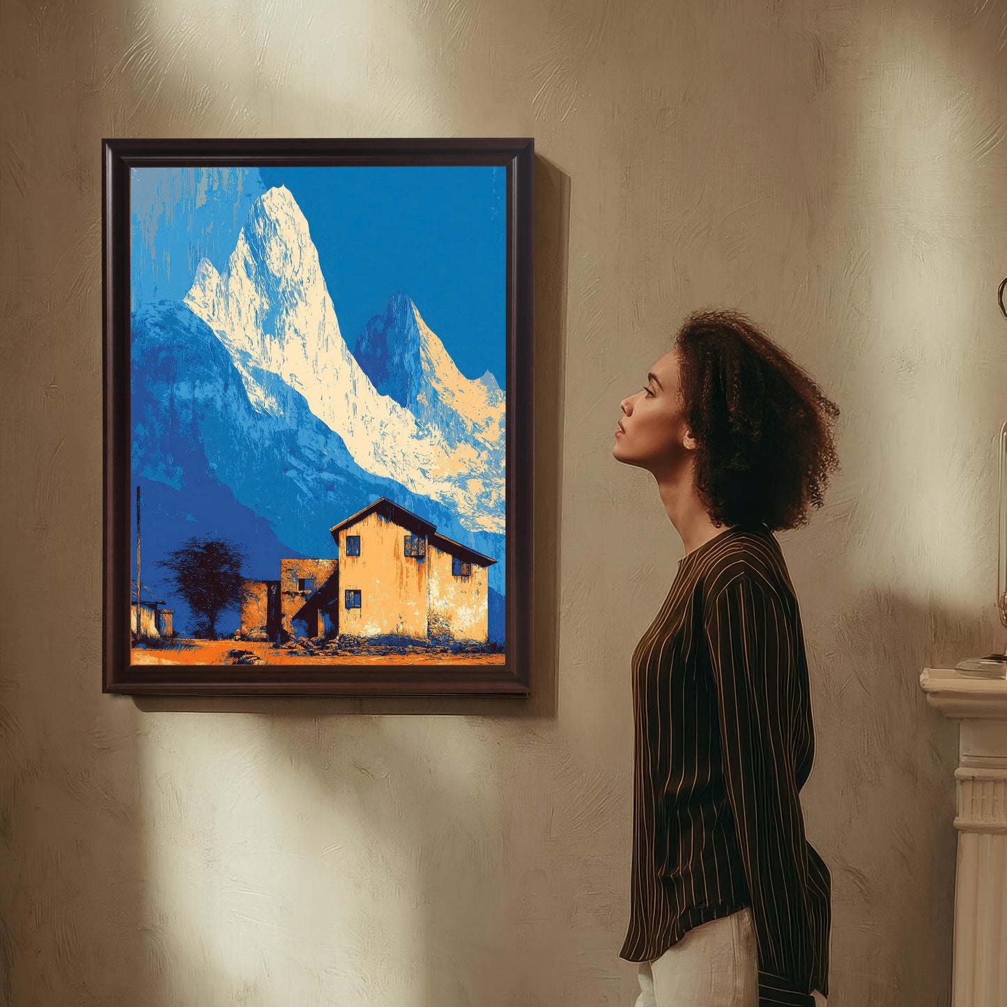 Framed art print with stylized white and blue mountains and an orange house, hanging on a textured wall, with a woman looking at the artwork.