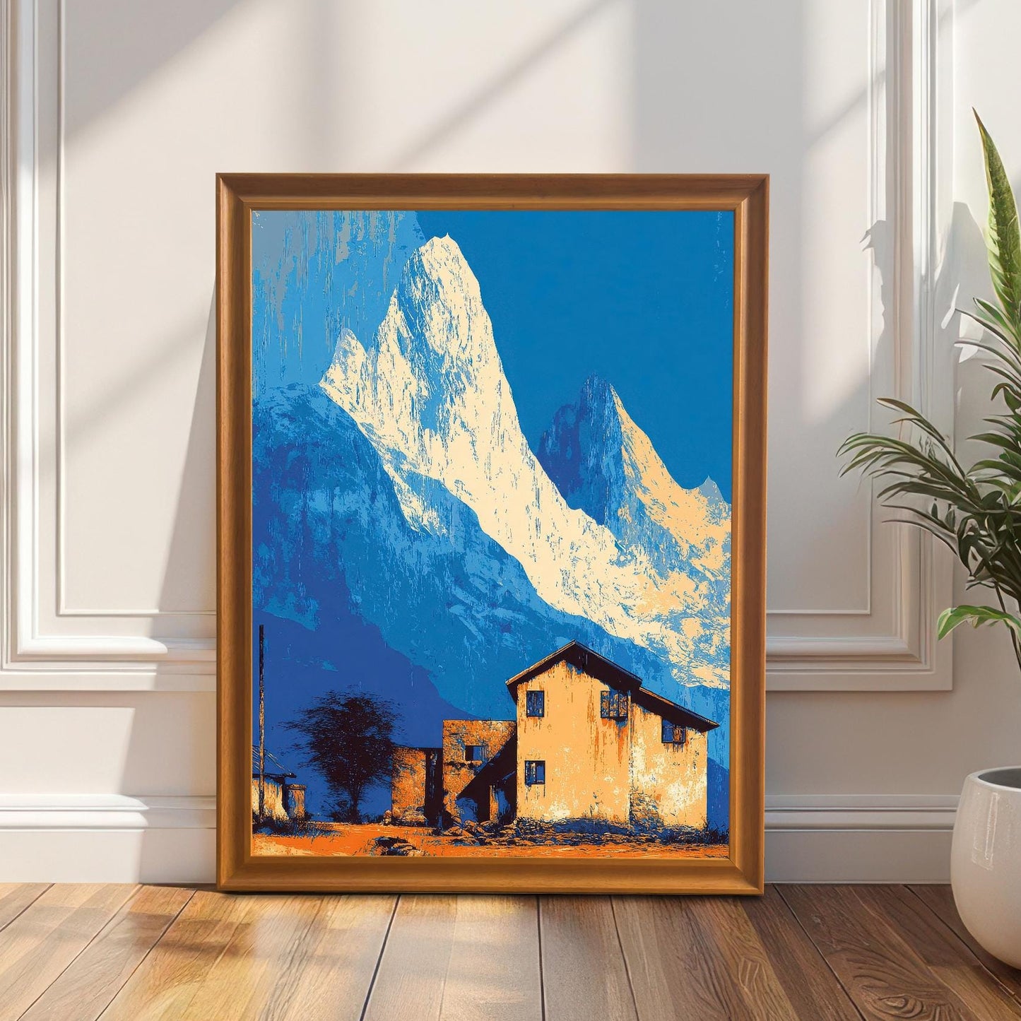 Landscape art print in an orange/light brown wood frame, featuring stylized blue mountains and an orange house, leaning on a white decorative wall next to a tall potted plant.