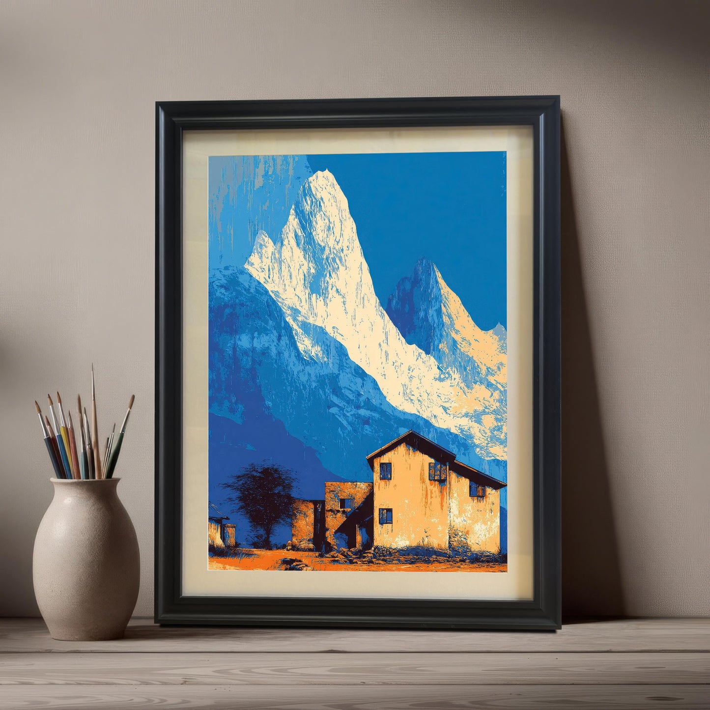 Landscape art print in a black frame with a mount, leaning on a shelf next to a vase of paintbrushes.