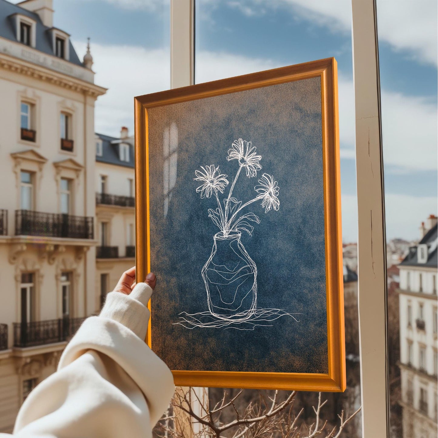 A hand holding the framed print in an orange/light brown wooden frame, held up near a window with a view of European-style apartment buildings outside. The art features a distressed blue and orange background.