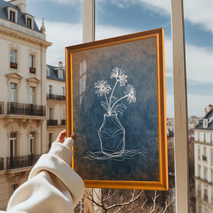 A hand holding the framed print in an orange/light brown wooden frame, held up near a window with a view of European-style apartment buildings outside. The art features a distressed blue and orange background.