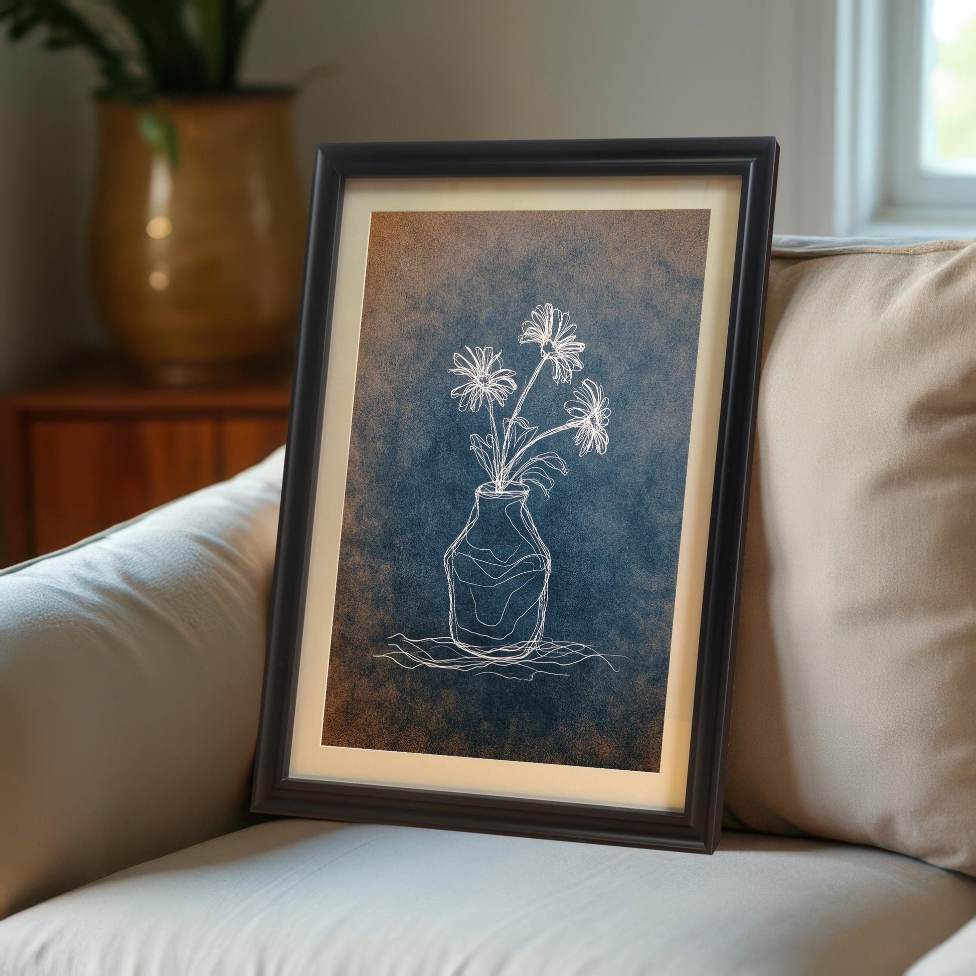 Close-up shot of the framed print resting on the arm or cushion of a cream-colored upholstered sofa. The print is in a dark brown/black frame with a mat and features a white flower drawing on a textured blue and orange background.