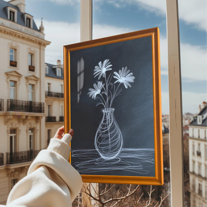 A hand holding the framed chalk flower print in an orange/light brown wooden frame, held up near a window with a view of Parisian-style apartment buildings outside.