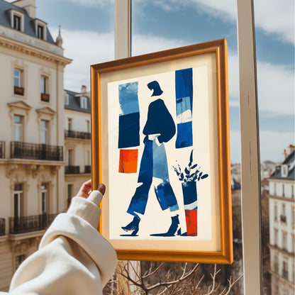 A hand holding the framed art print in an orange/light brown wooden frame, held up near a window with a view of European-style apartment buildings outside.