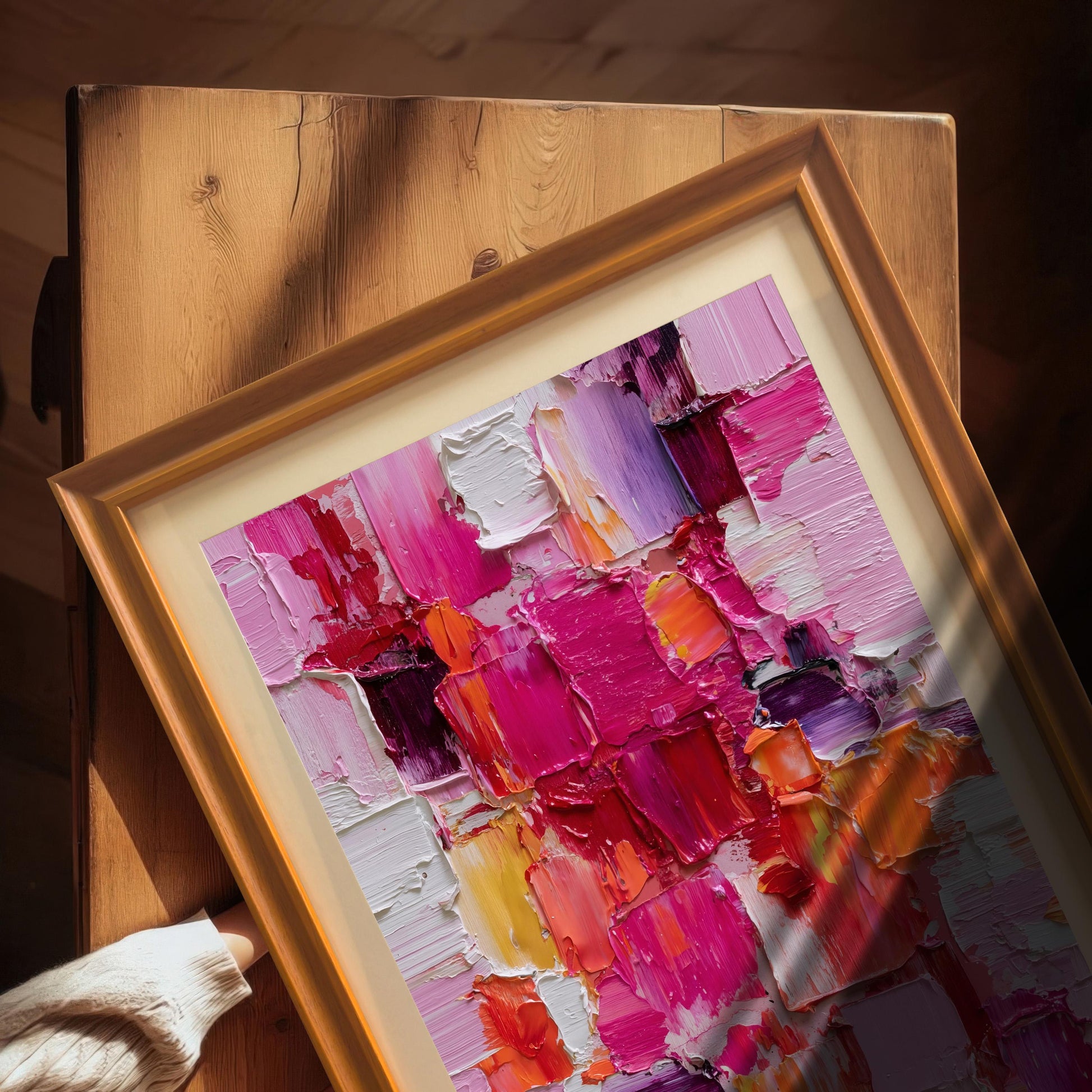 Abstract oil-style painting in an Orange wooden frame with an off-white mat, resting on a rustic wooden table with bright sunlight casting shadows. A person's hand in a white sweater is near the bottom edge.