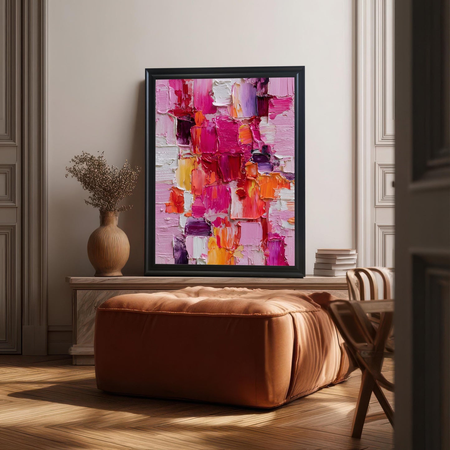 Abstract oil-style painting in a black wooden frame without a mat, resting on a console table or mantelpiece behind a large, square, camel-brown leather ottoman/pouf. The room has light wood flooring and classical white wall paneling.