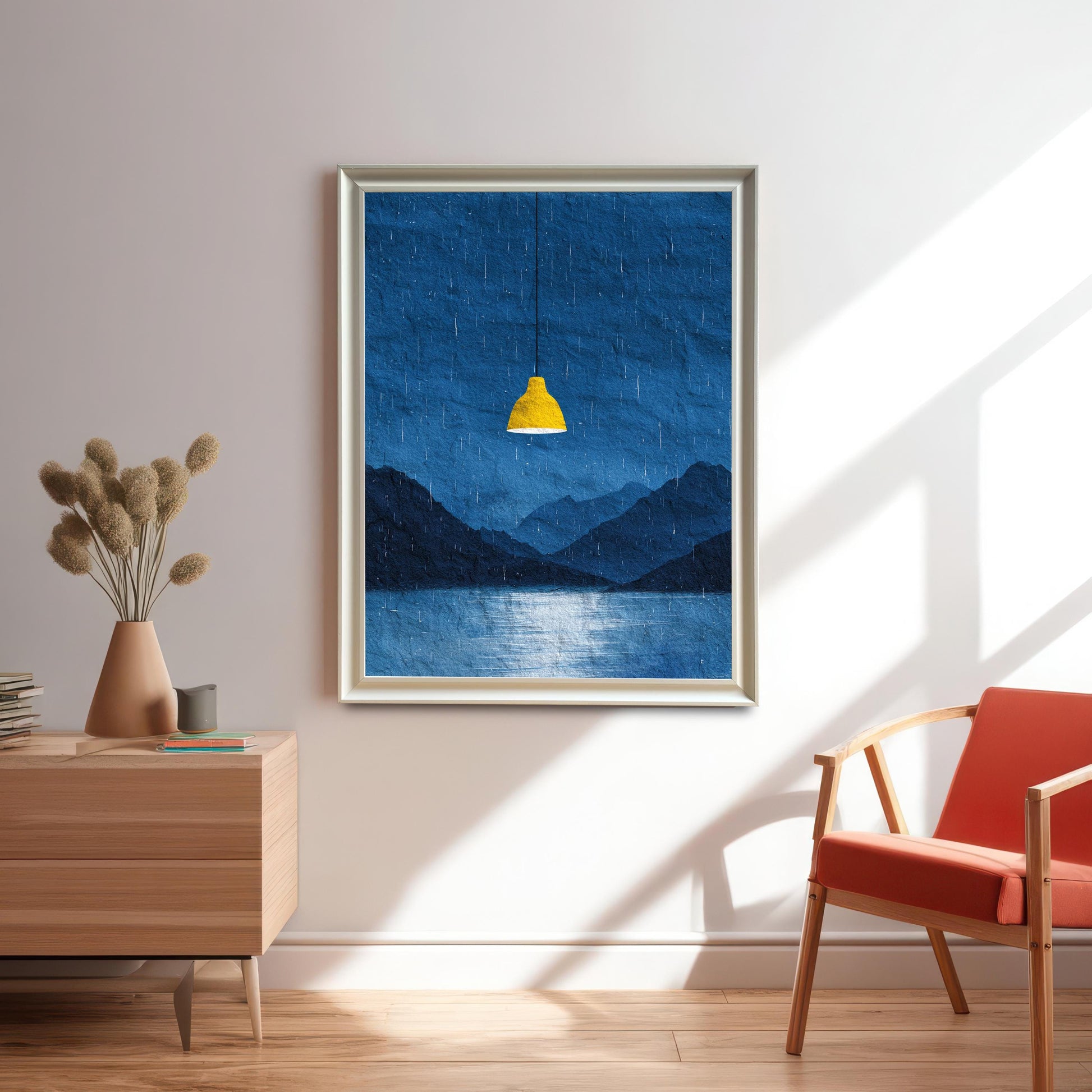 A framed art print of a dark blue, rainy mountain and lake landscape with a bright yellow hanging lamp. The print is in a white/light grey frame, hanging on a white wall above a wooden cabinet. A terracotta vase with dried flowers and an orange chair are also in the sunny room.