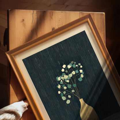 Overhead close-up view of the golden vase and eucalyptus art print in an Orange frame with a mount, resting on a wooden surface. A hand in a cream sweater is visible in the corner.