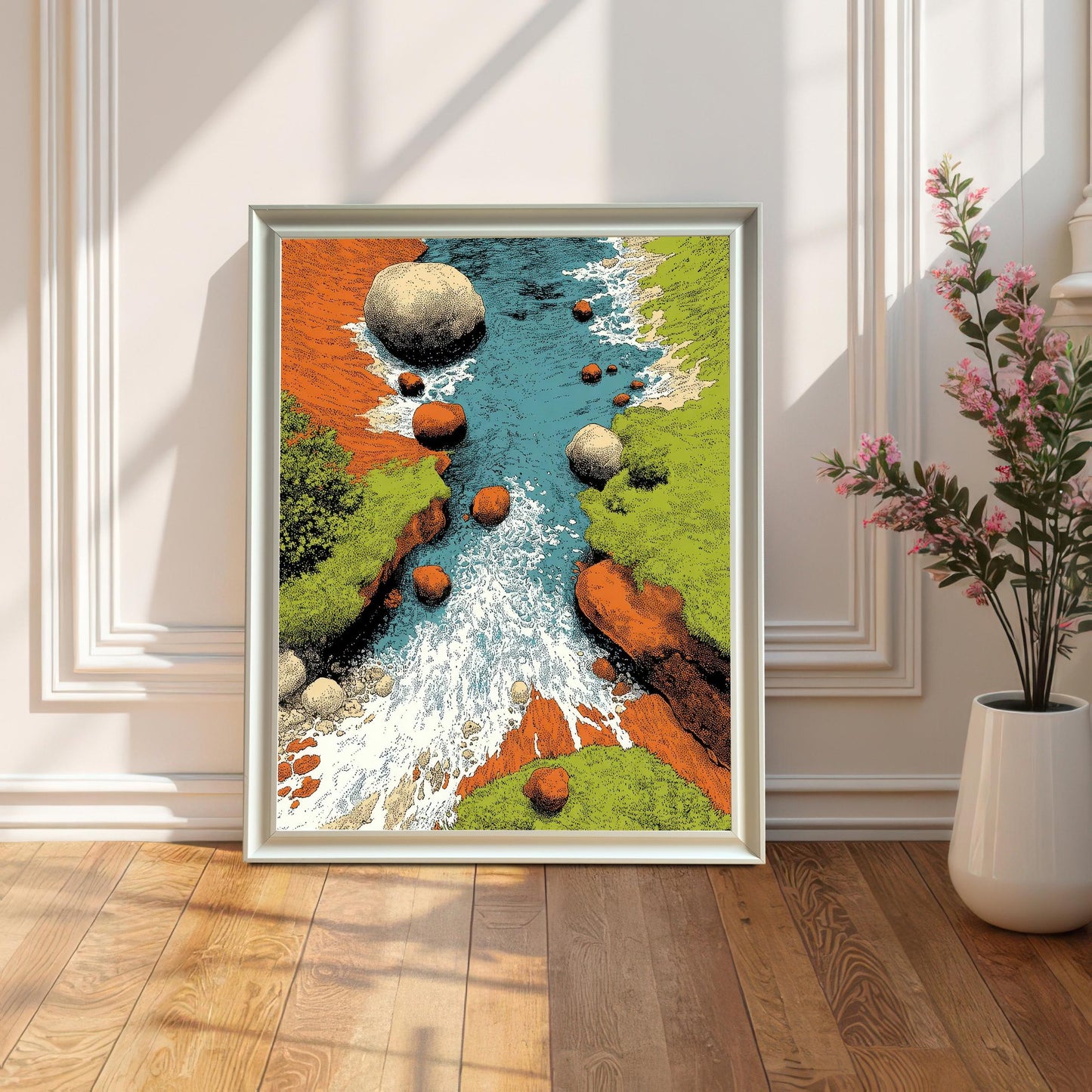 A framed art print in a white frame, leaning on a wooden floor against a white paneled wall (wainscoting). The artwork is a colorful, stylized illustration of a river rushing between rocky, green banks. Sunlight casts long, dramatic shadows. A large white vase with pink flowers sits to the right.