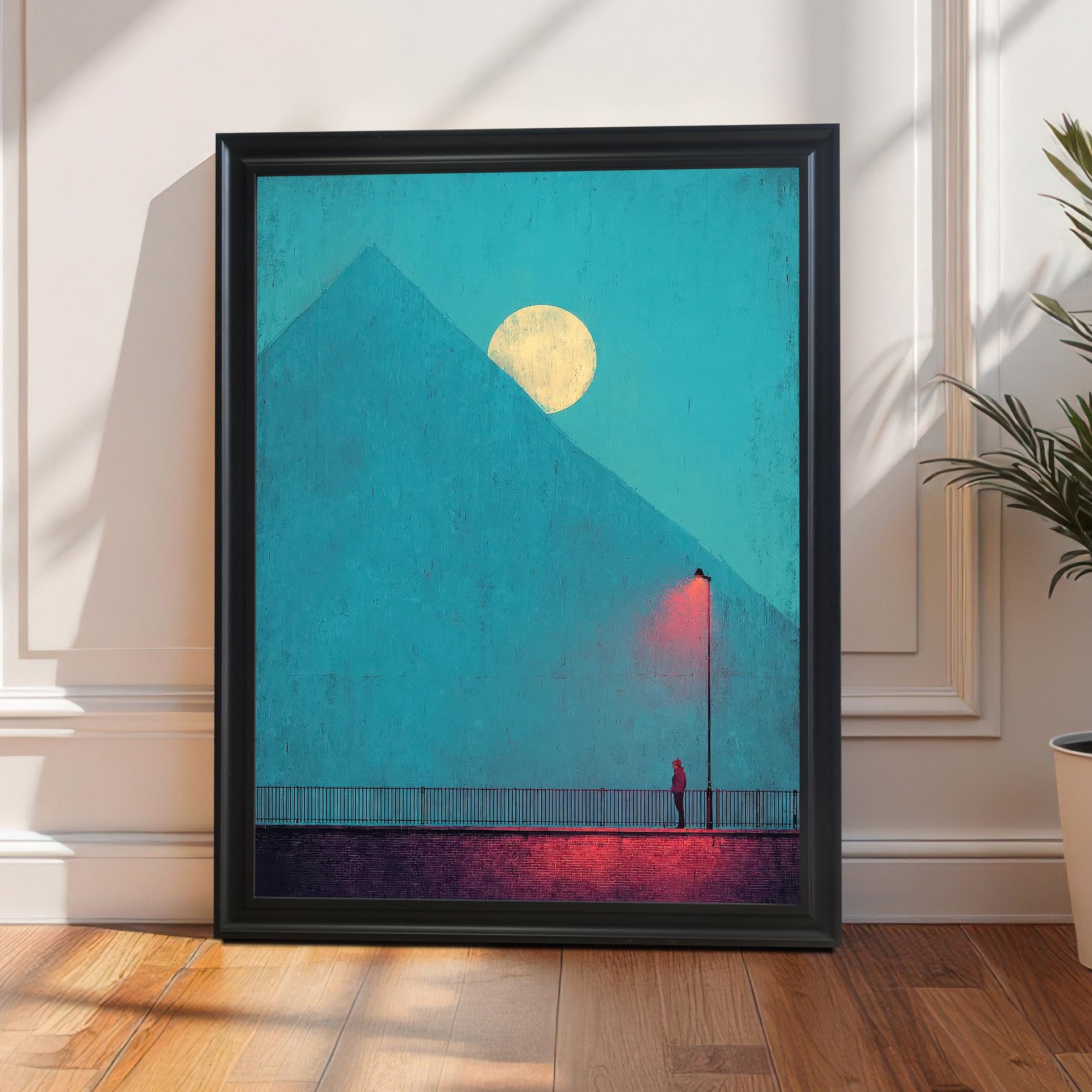 A framed art print in a black frame, leaning on a wooden floor against a white paneled wall (wainscoting). The artwork is a moody, minimalistic scene with a figure under a red lamp post and a yellow moon behind a blue pyramid/mountain. Sunlight casts long, dramatic shadows. A potted palm or similar plant sits to the right.