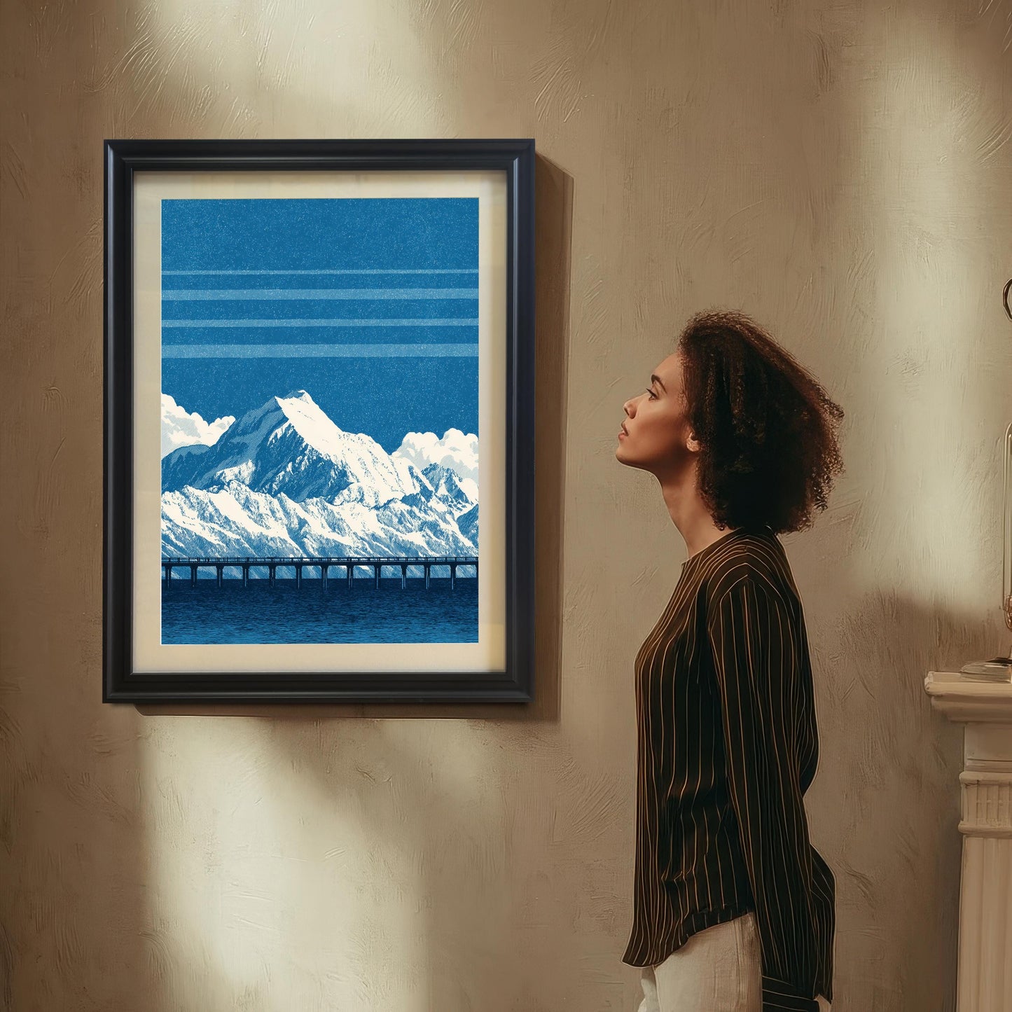 A woman in a brown striped shirt looks up at a framed art print of a blue mountain and bridge scene hanging on a textured beige wall. The print is in a black frame with a light-colored mount/mat.